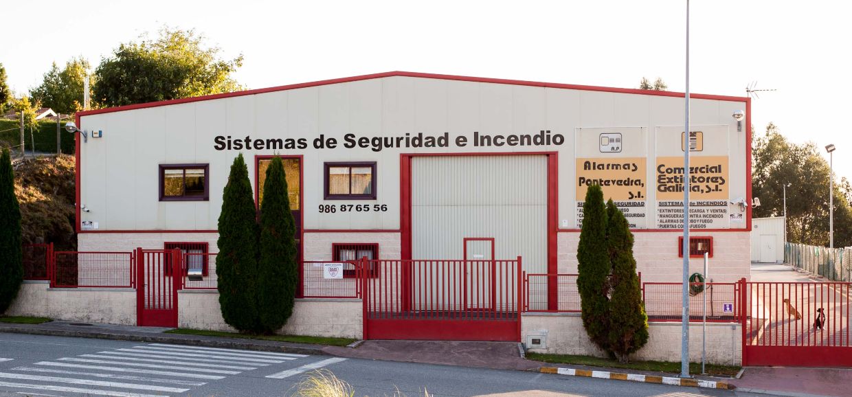 Frontal de la nave de la empresa Comercial Extintores Galicia, S.L. con fachada blanca y portales rojos rodeada de árboles